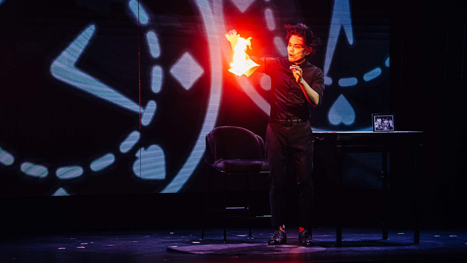 Shin Lim on stage standing in front of a chair and table while dropping something that is on fire.