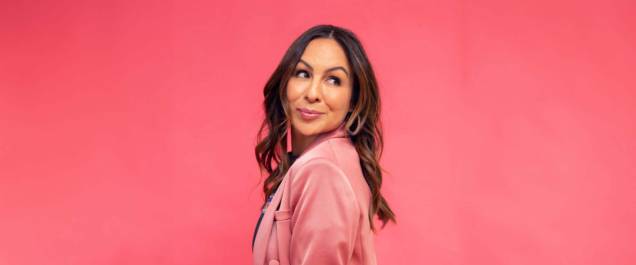 Anjelah Johnson-Reyes in a pink jacket, looking over her shoulder, with a pink backdrop