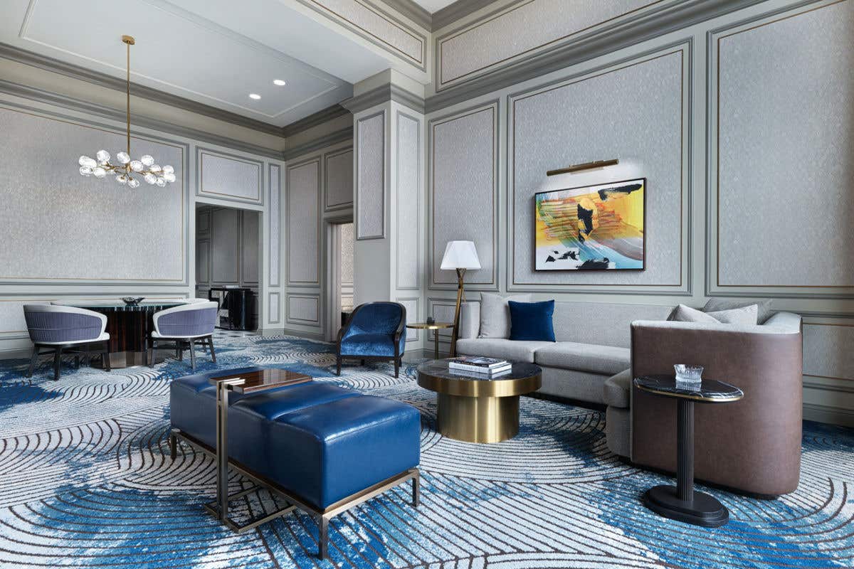 A spacious, modern living room featuring a gray sectional sofa, blue lounge chairs, a round gold coffee table, and abstract artwork on paneled walls, with patterned blue carpeting and a small dining area in the background.