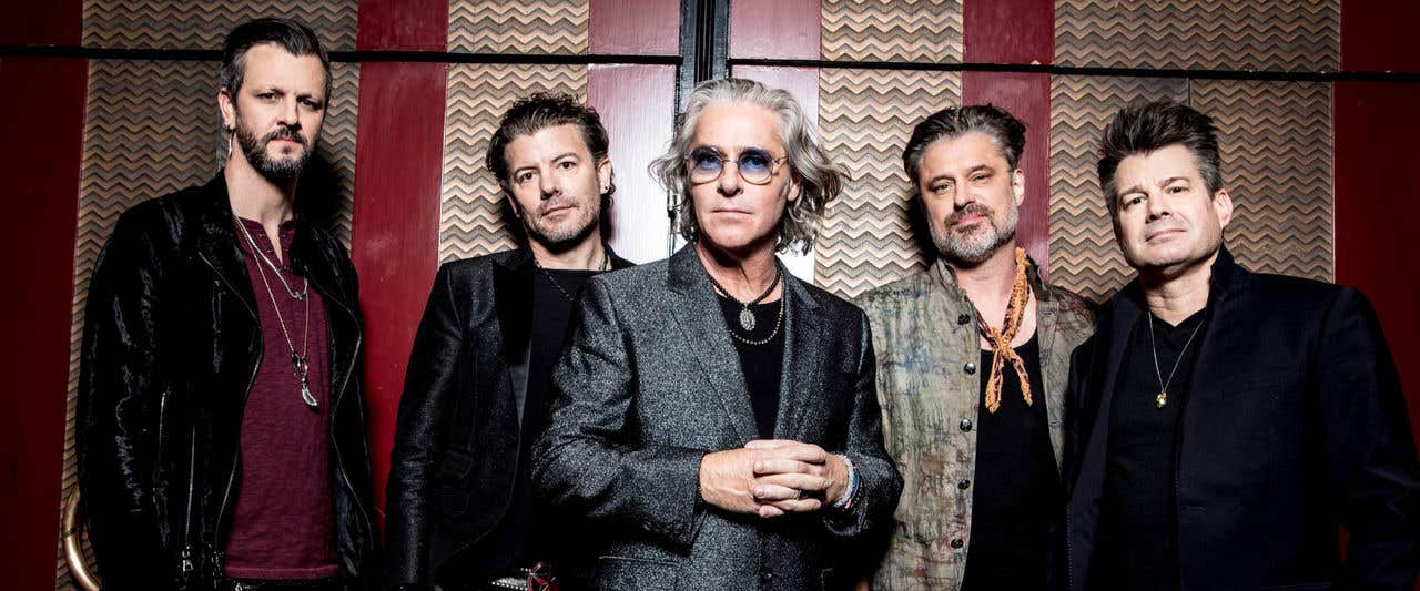 Collective Soul members standing in front of red and tan striped patterned wall staring at the camera with Ed Roland front and center. 