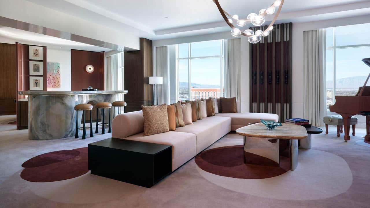 A large hotel living room during the day with a couch, coffee table, baby grand piano, bar seating, and large windows with a view of the Las Vegas Strip.