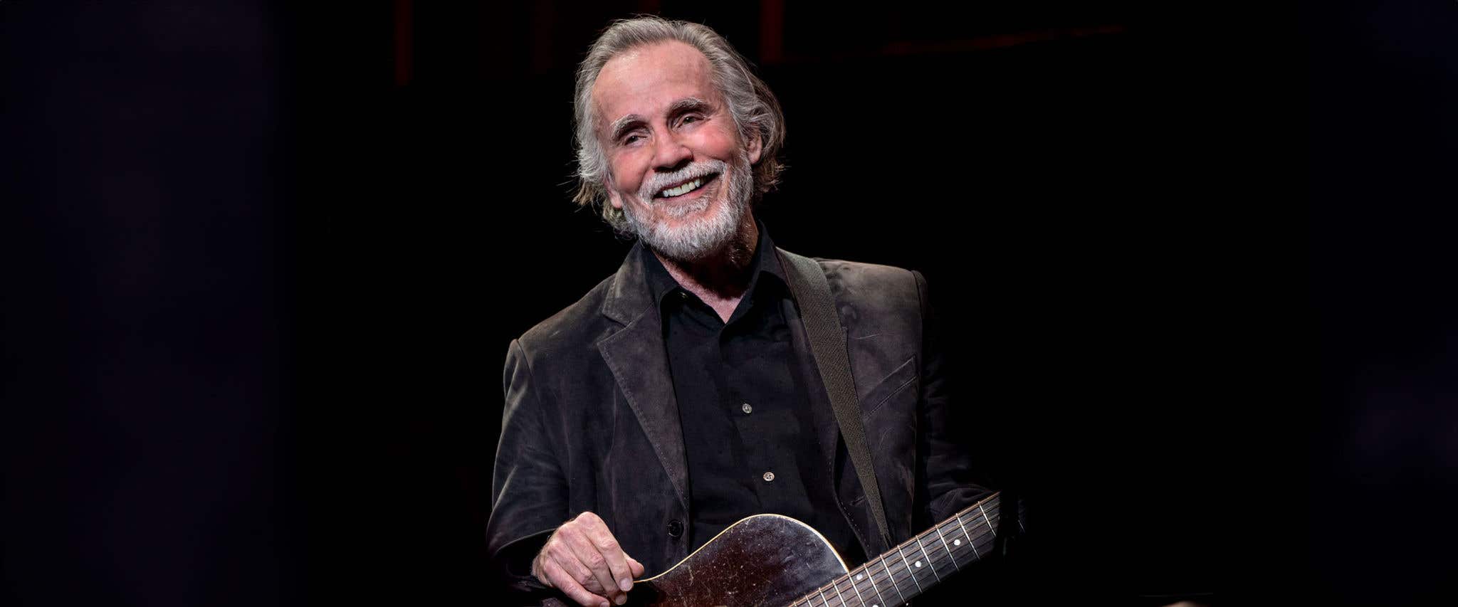 Jackson Browne, in dark attire, smiling and holding a guitar against a black backdrop
