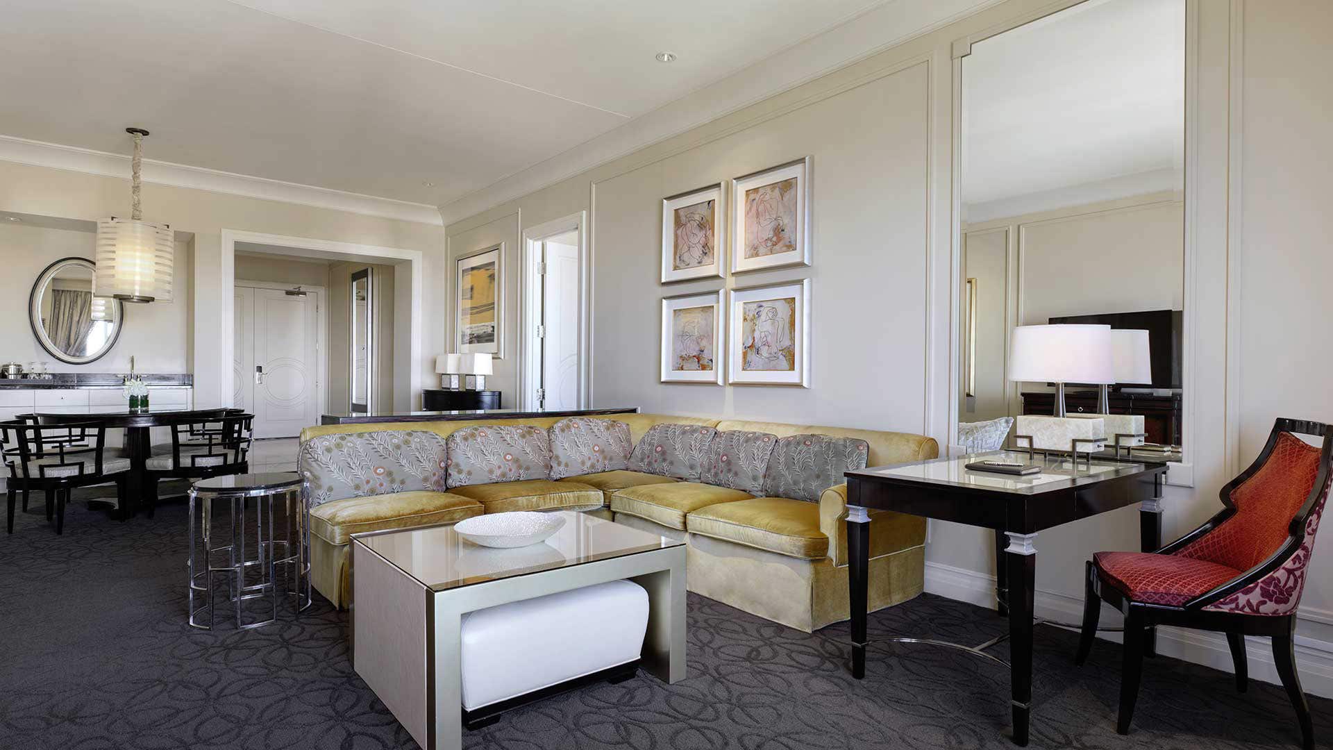 A hotel room living room with an L-shaped couch, coffee table, desk, and four-seater dining table behind it.