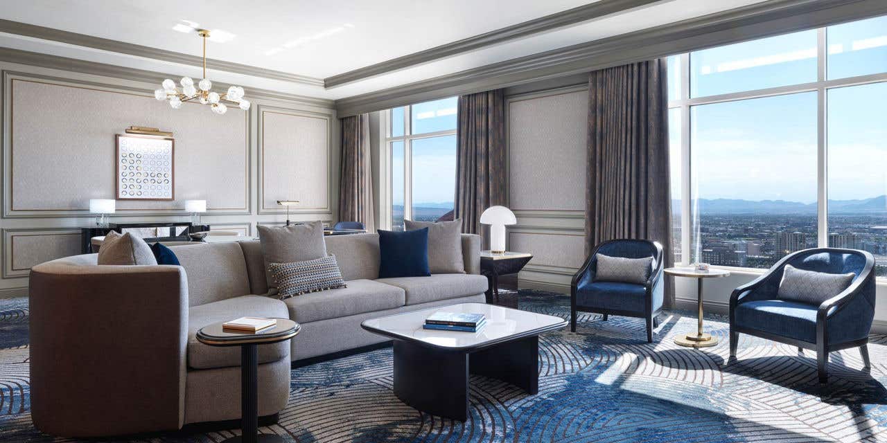 A hotel living room with a couch, coffee table, two armchairs, a dining table, a desk, and two large windows with a view of the Las Vegas valley.