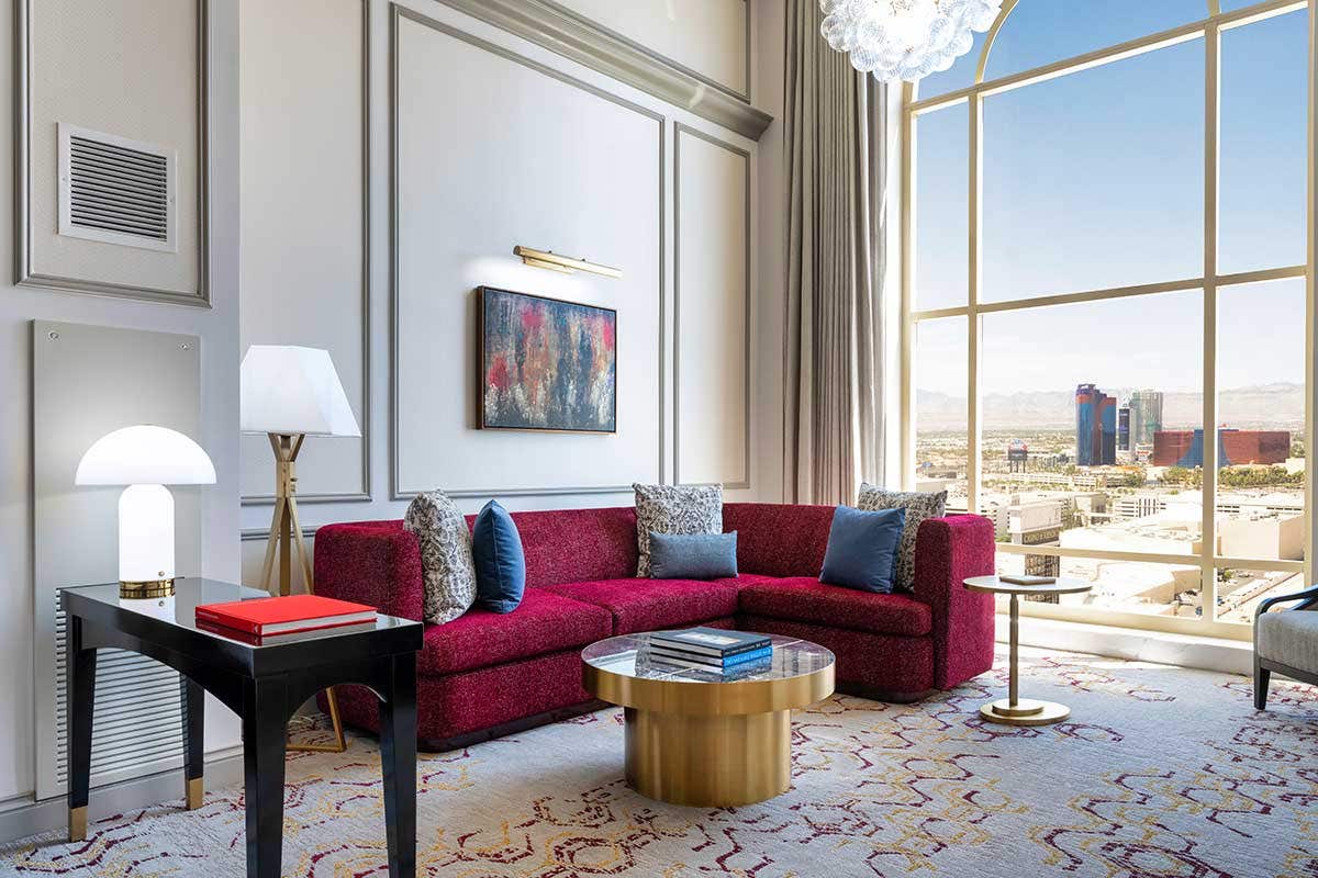 A hotel living room with a side table, L-shaped red couch, gold coffee table, and large window on the right.