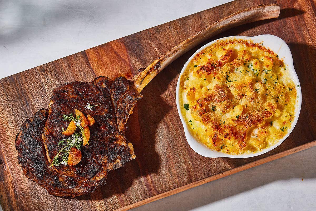 A grilled tomahawk steak and a serving of macaroni and cheese sit on a wooden cutting board.