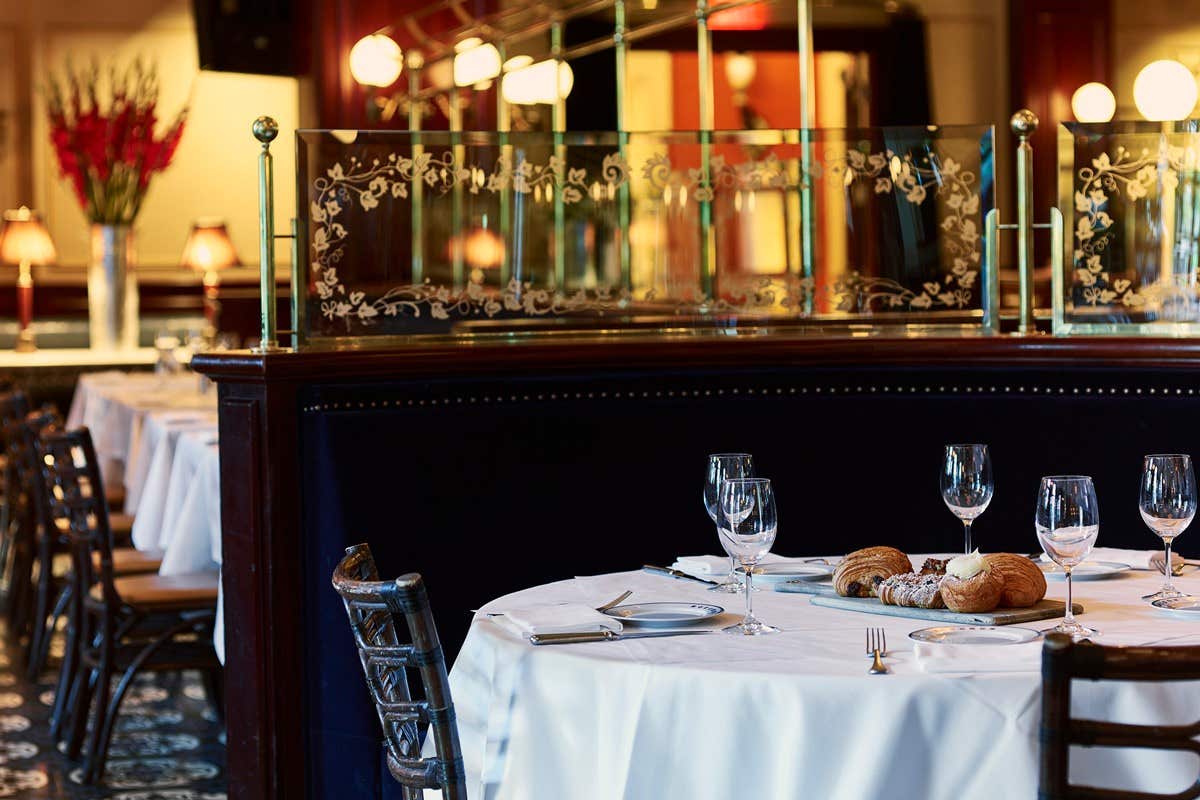 Elegant restaurant with a set table, empty wine glasses, and bread rolls. Warm lighting and a decorative divider in the background.