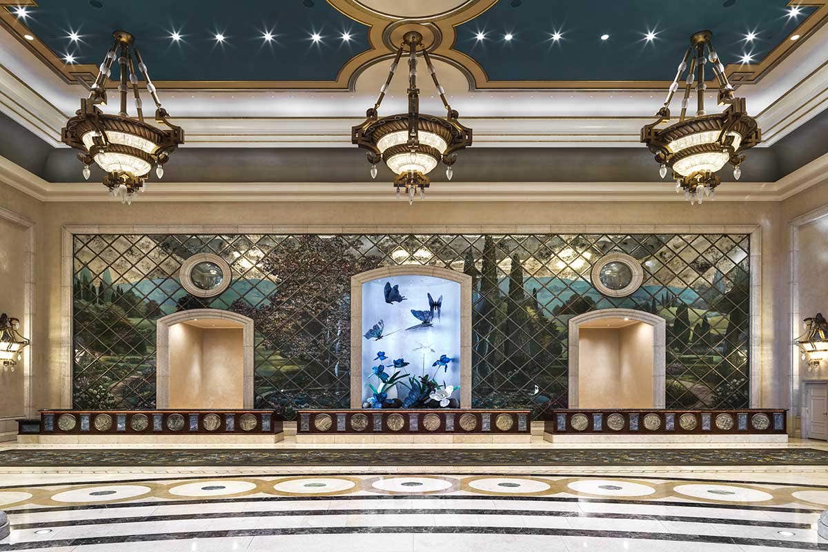 A spacious hotel lobby with three chandeliers and a long check-in desk, with a display of large blue butterflies behind it.