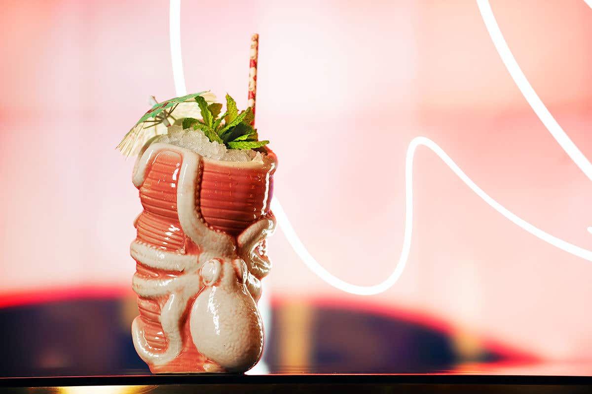 A pink colored ceramic mug with a decorative octopus on the outside, containing ice, mint and a paper umbrella, against a pink colored backdrop and a wavy light stream