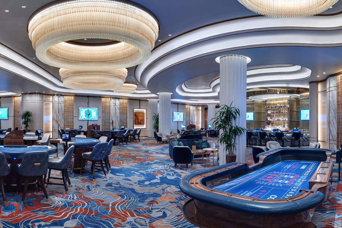 A spacious, upscale casino floor featuring gaming tables, plush seating, large sculptural chandeliers, curved architectural ceilings, and vibrant patterned carpeting, with a bar area and multiple screens visible in the background.