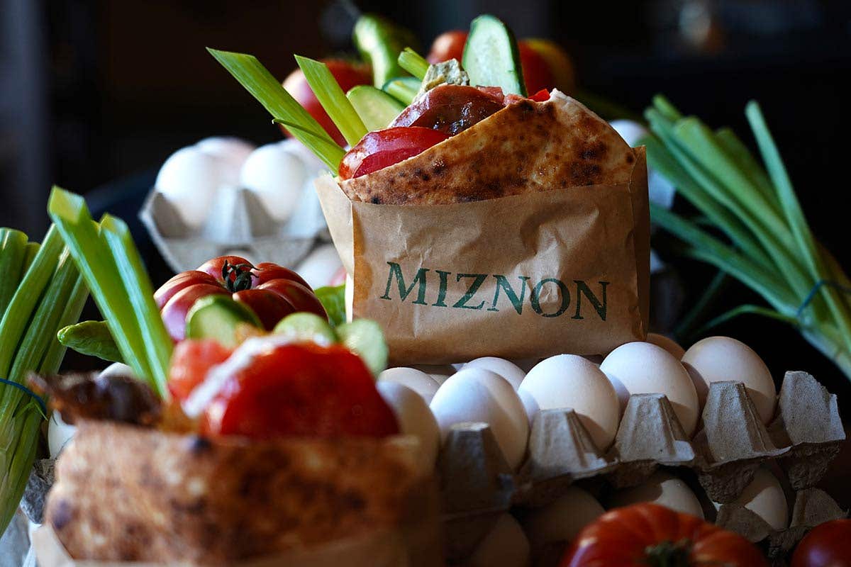 Pita sandwich in a Miznon paper wrap sitting on top of a carton of eggs and surrounded by green onions and tomatoes