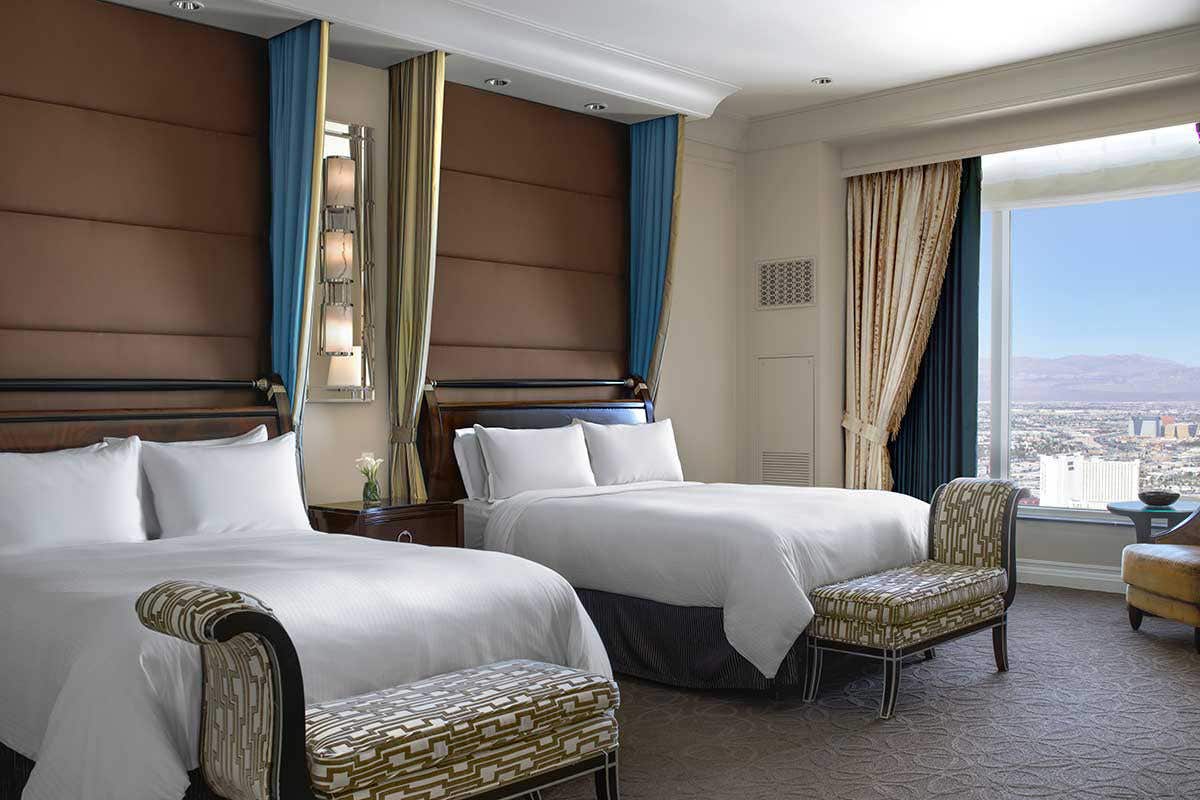 A hotel room with two Queen beds, bench at the end of each bed, and a large window to the right overlooking the city and mountains.