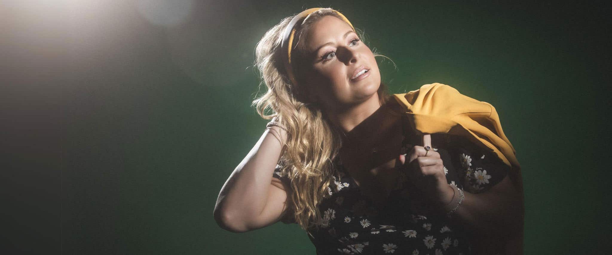 Becky Robinson in a black floral top with a yellow headband and jacket against a dark green background.