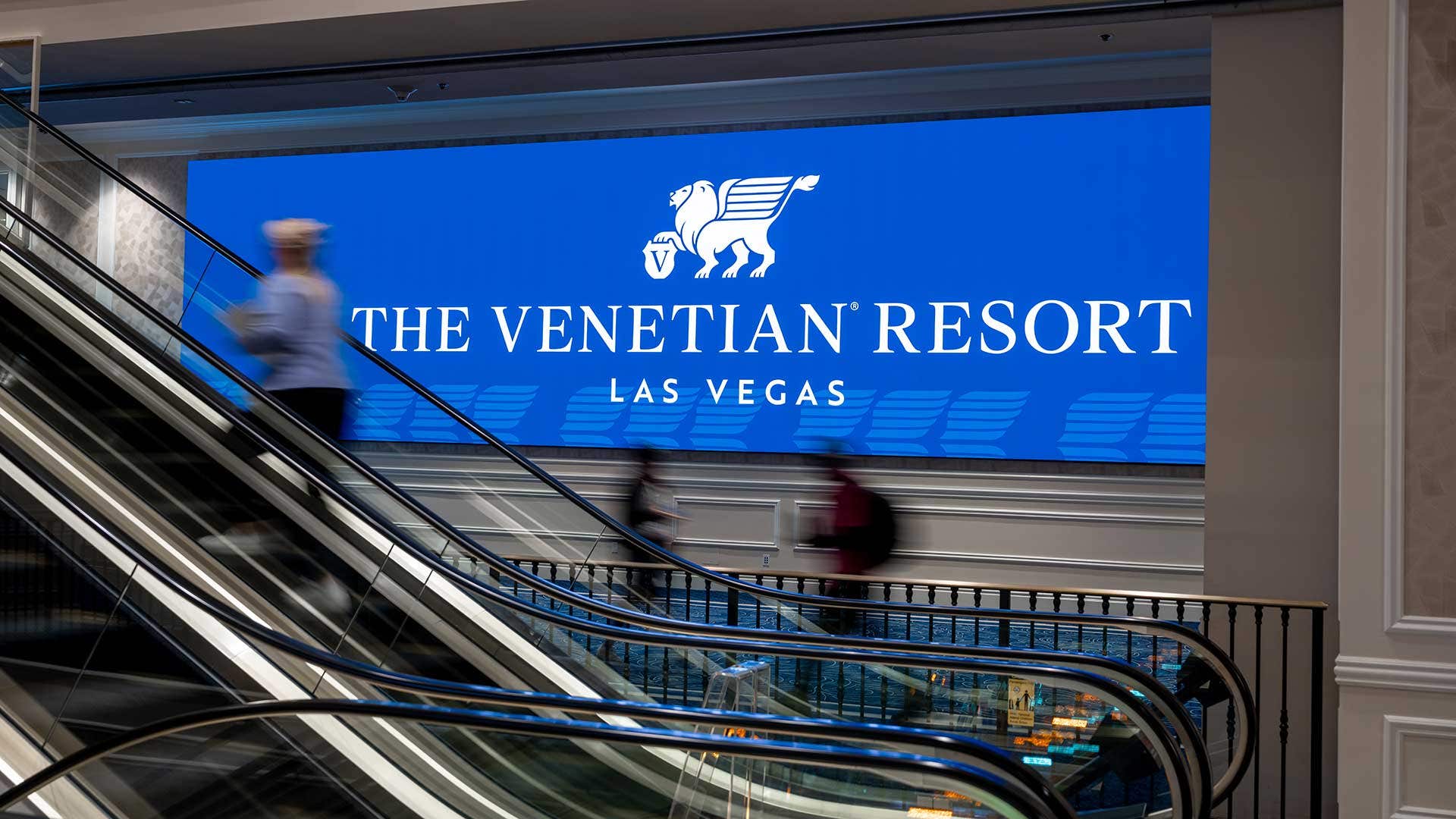 Large digital screen on the wall by a big hallway with blue carpet in front of escalators.