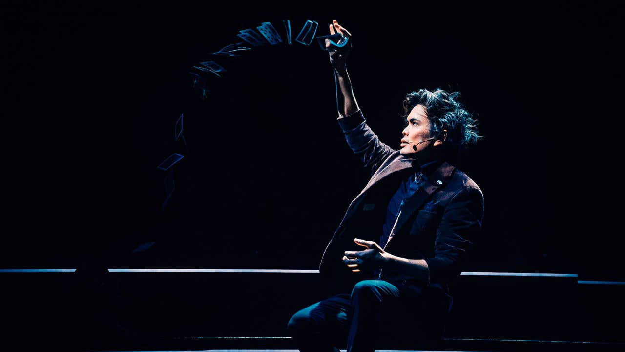 Shin Lim sitting on stage looking at his hand release a deck of cards into the air.
