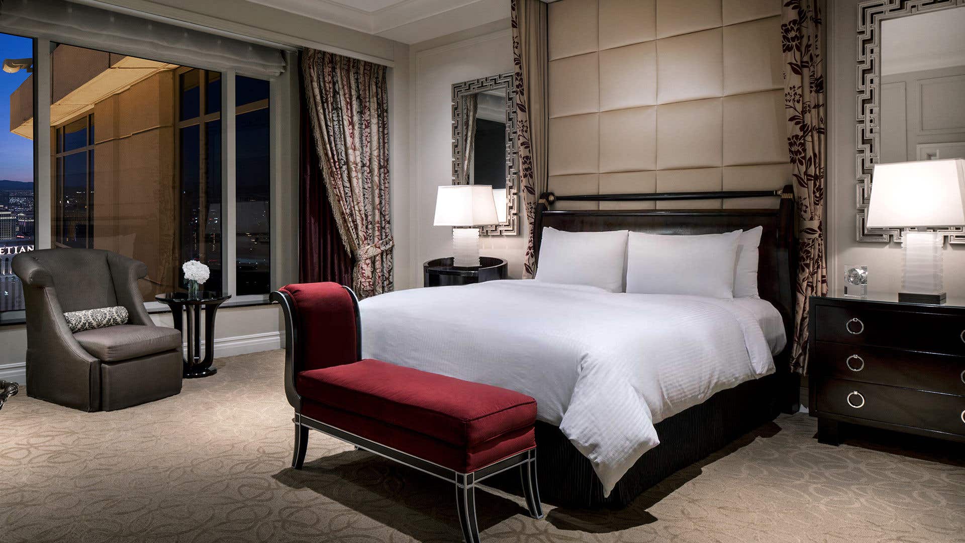 Hotel room at night with a king-sized bed with white linens and a plush headboard, flanked by nightstands with lamps and a red bench at the foot of the bed. A window with a city view on the left with an armchair and side table in front of it.  