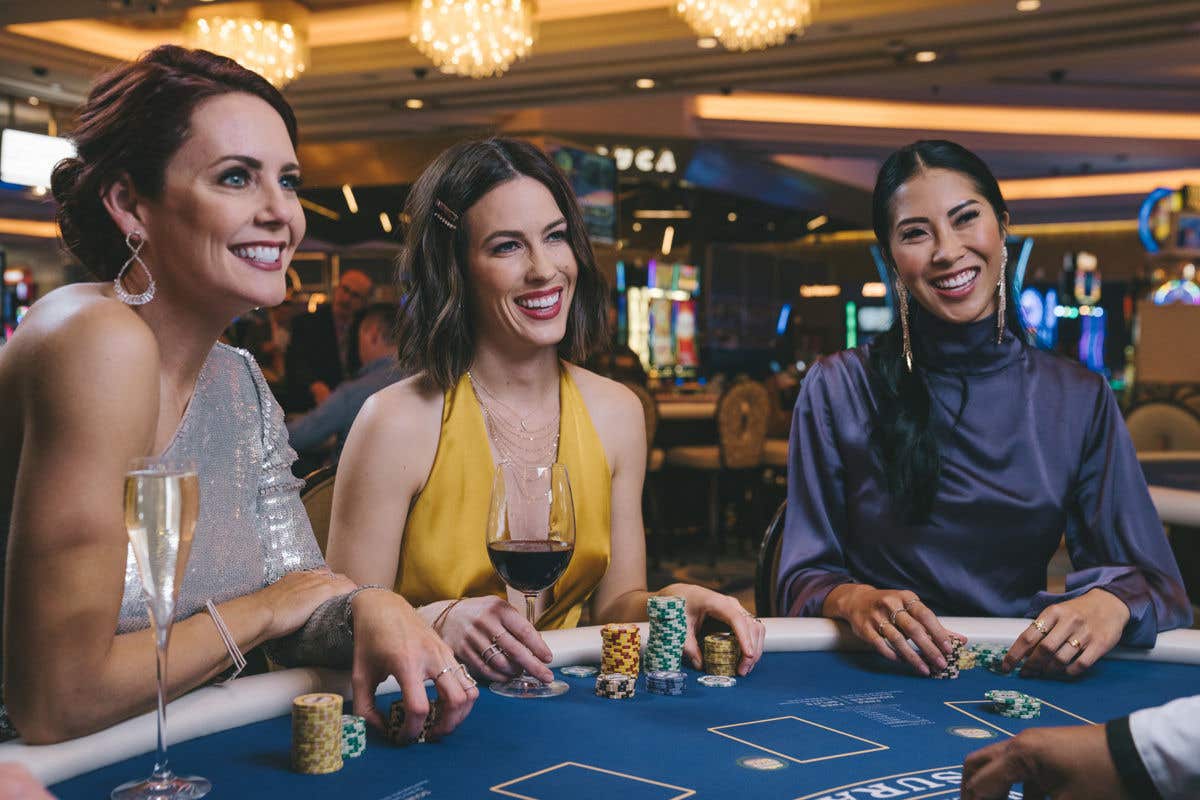 Three people dressed in evening attire sit at a casino table, holding chips and drinks while playing a game under warm overhead lighting.