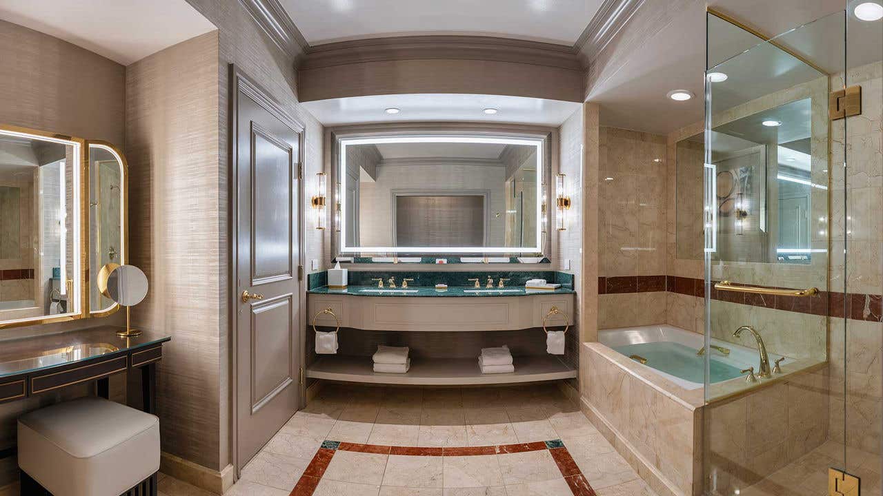 A hotel room bathroom with a lighted mirror, two sinks, a bathtub and separate shower on the right, and a makeup vanity and door on the left.