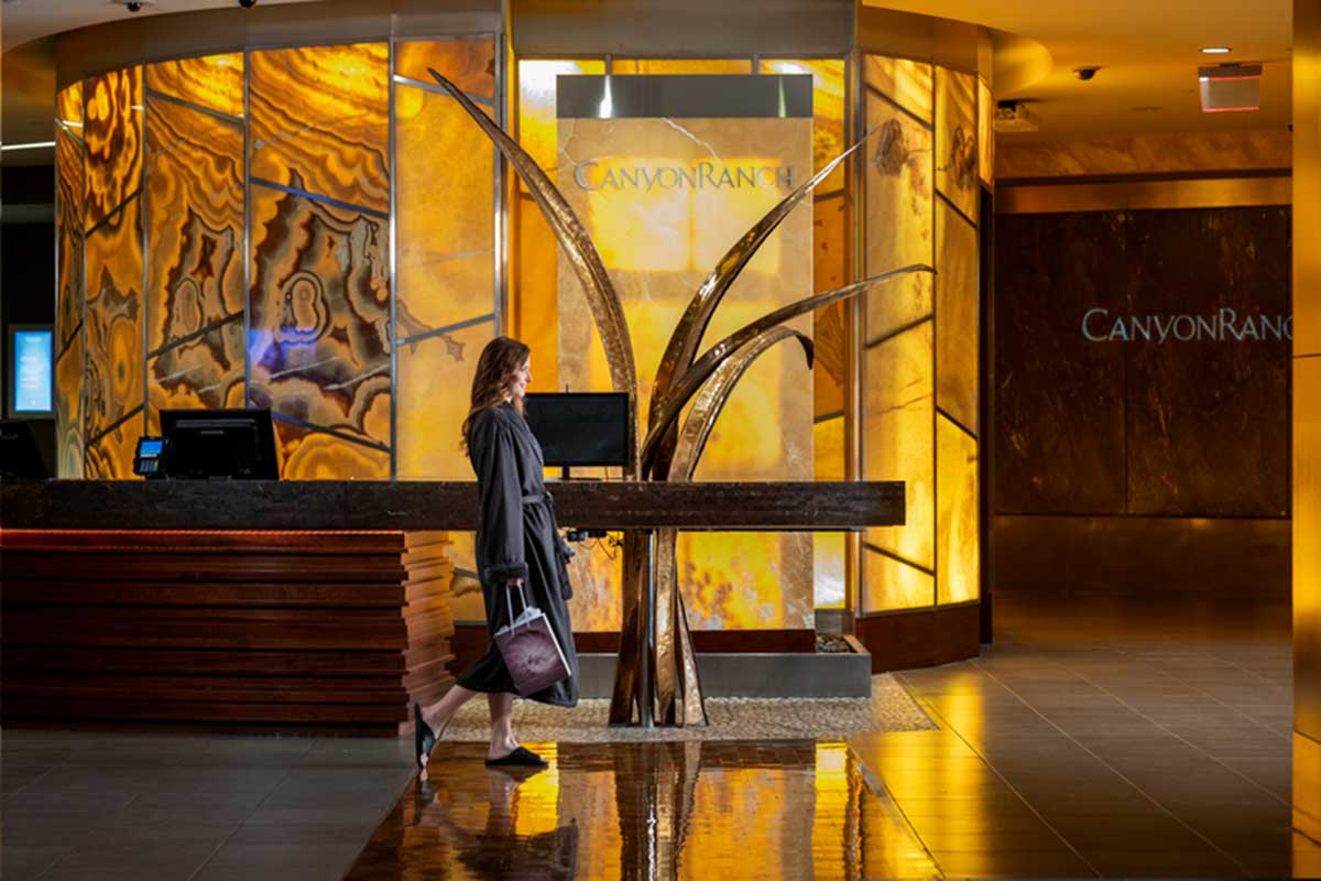 Canyon Ranch Spa and Fitness lobby with a check-in desk to the left and a woman in a robe walking to the treatment rooms on the right.