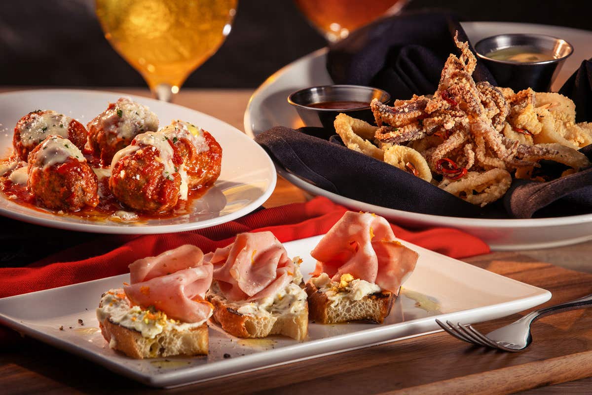 Plates of Italian meatballs, fried calamari, and bruschetta with cured meat and cheese on a wooden table with red napkins.