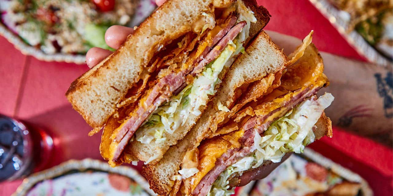 A hand holding a large sandwich with meat, chips, and lettuce above a red table. 