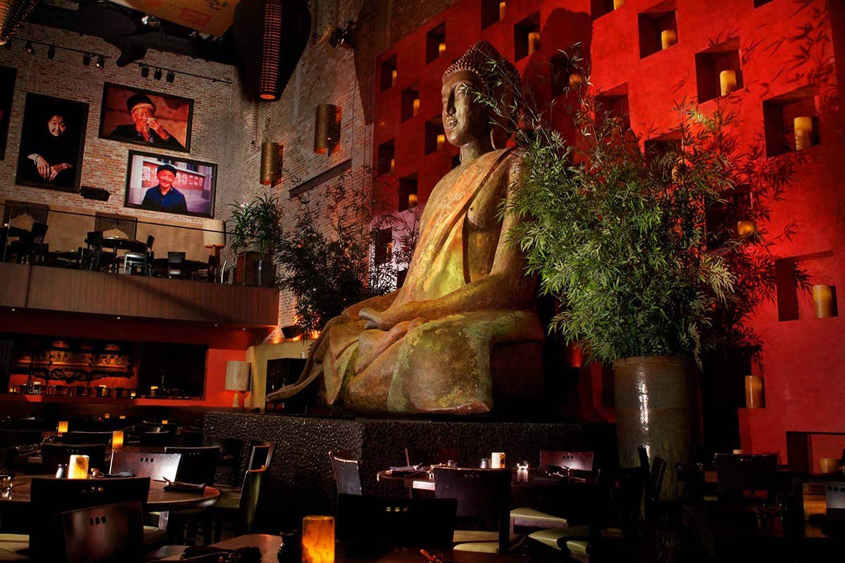 A large Buddha statue in a dimly lit restaurant with red walls and candles, greenery surrounding the statue.