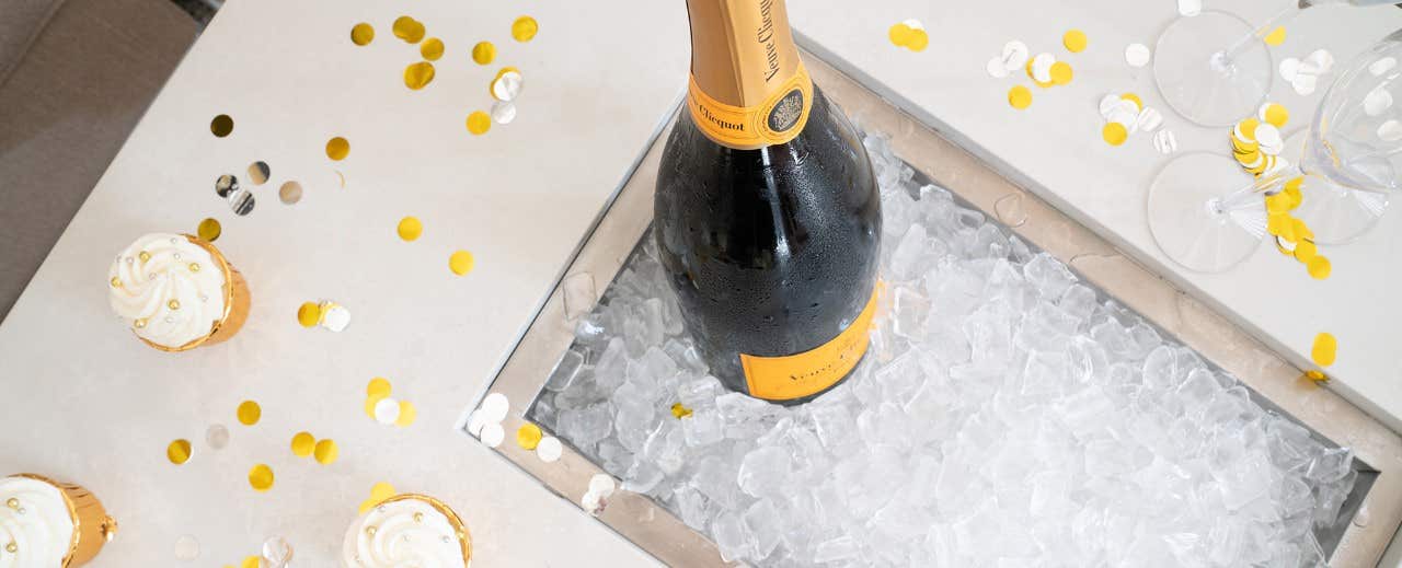 A bottle of champagne chilling in ice, surrounded by gold confetti, with three cupcakes and two empty champagne glasses.