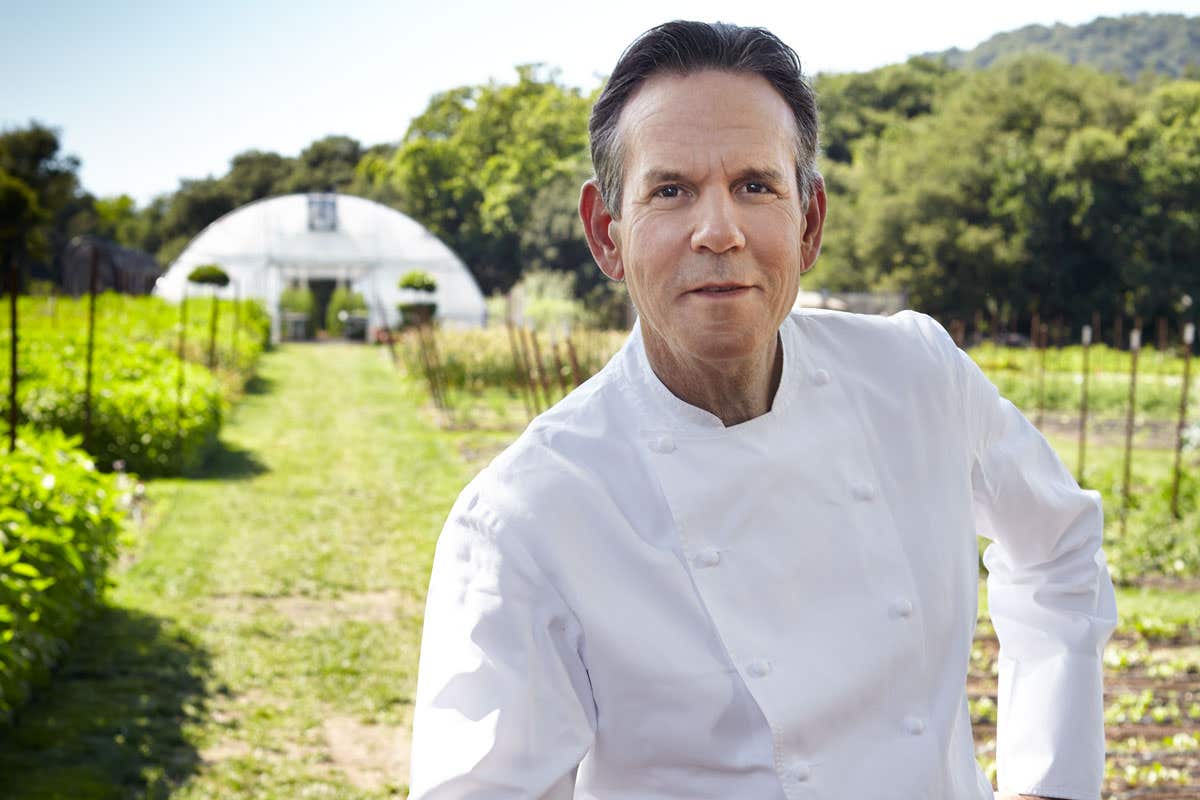A chef in a white coat stands outdoors in a garden with a greenhouse in the background on a sunny day.