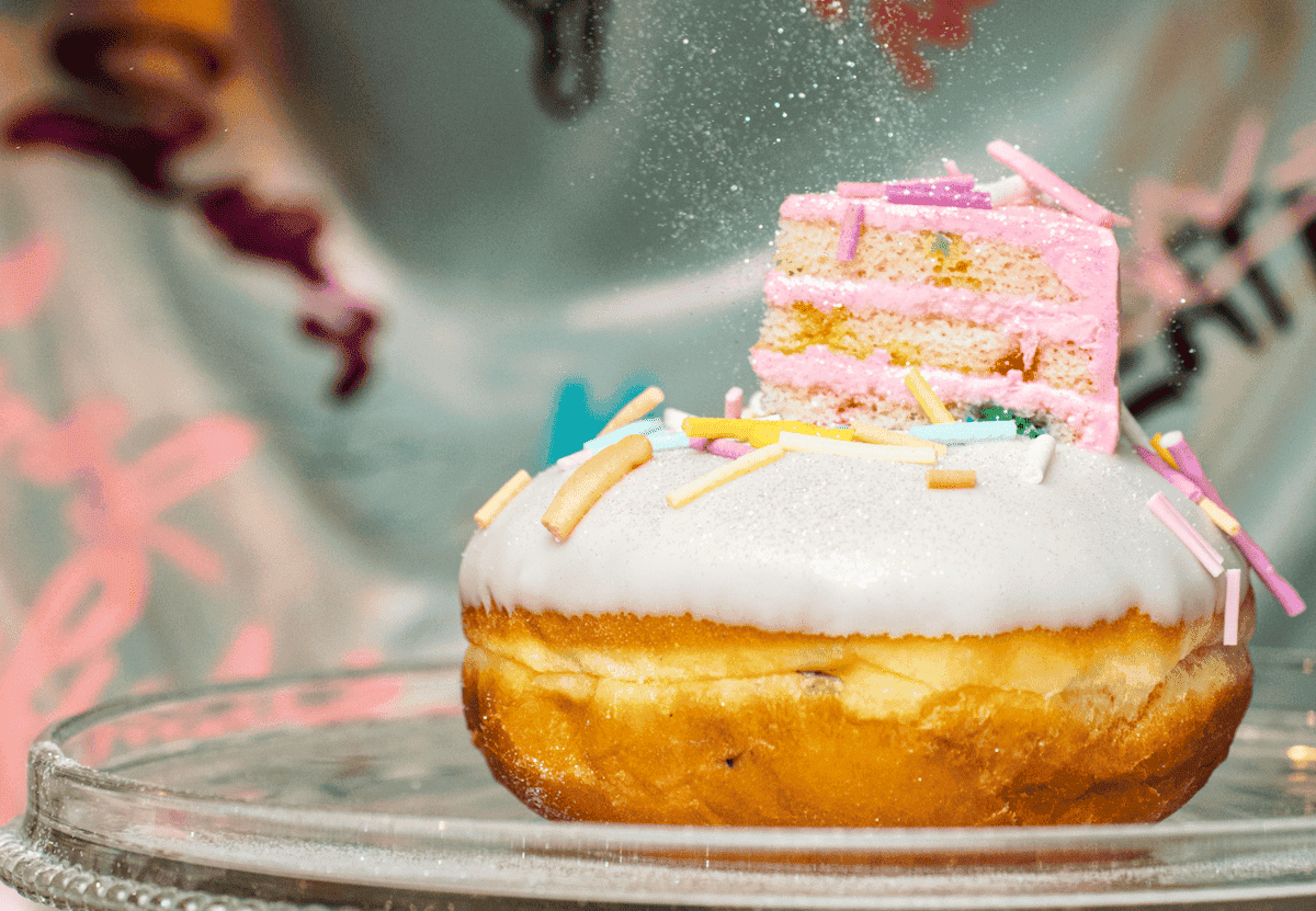 White donut with white frosting and sprinkles garnished with a mini slice of cake and icing sugar