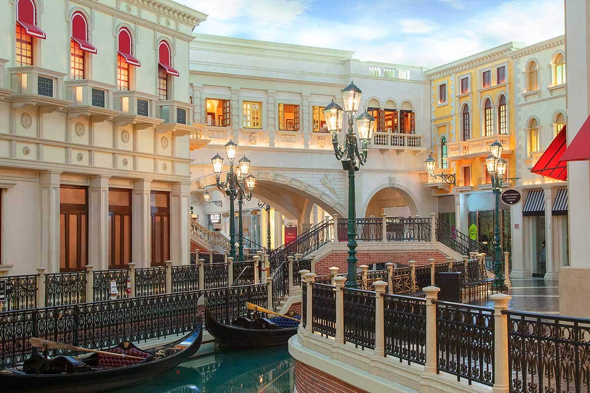 A view down an old-world looking setting of a canal with storefronts lined up against it with two gondolas sitting in the water between two lampposts