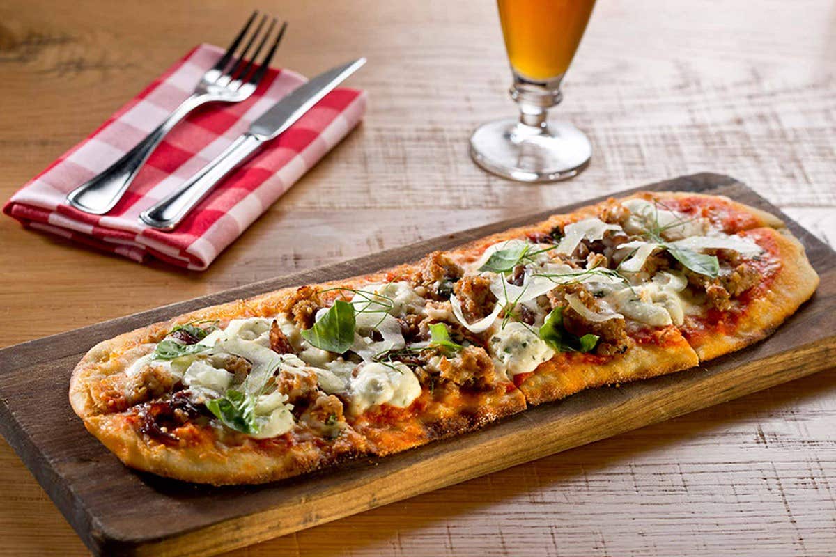 Long, rectangular pizza with various toppings on a wooden board, fork and knife with napkin, and a glass of beer on a table.