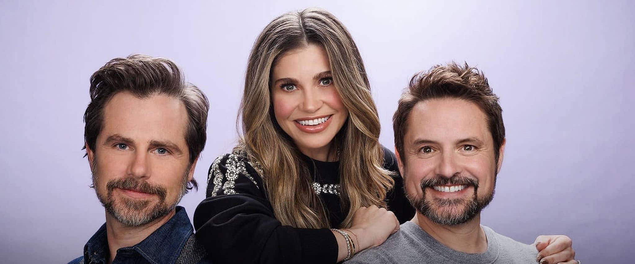 A group of three people, two men on the edge of the photo, and a woman in the center posed against a light purple colored backdrop