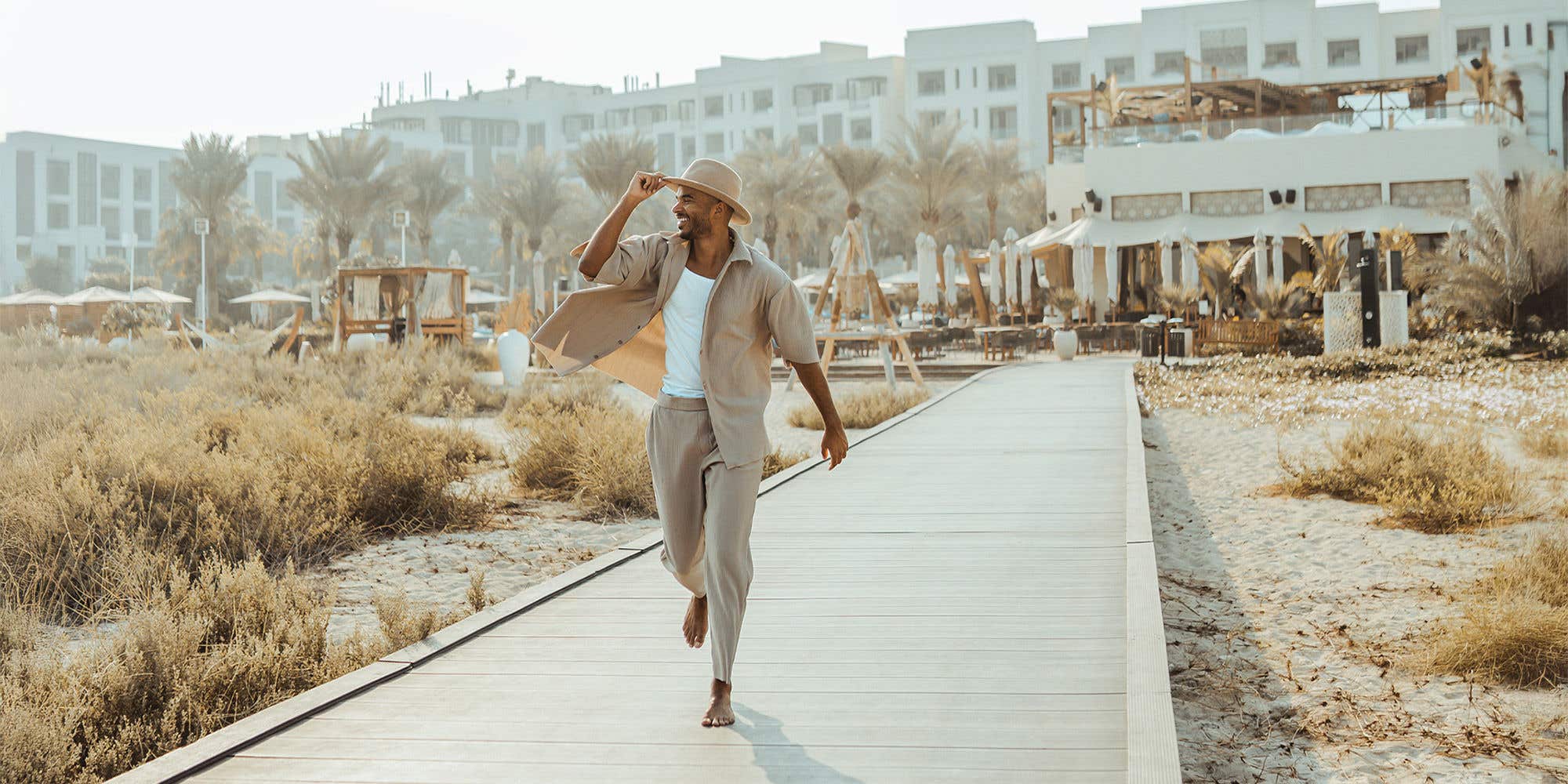 A man walks barefoot on a beach boardwalk, smiling and holding his hat, dressed in light casual clothes with a resort, palm trees, and sandy dunes in the background.