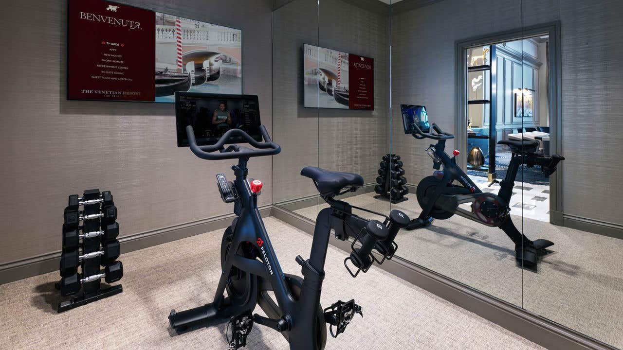 A workout room with a Peloton Bike facing a wall-mounted TV, a rack of dumbbells on the floor, and mirrored wall on the right.