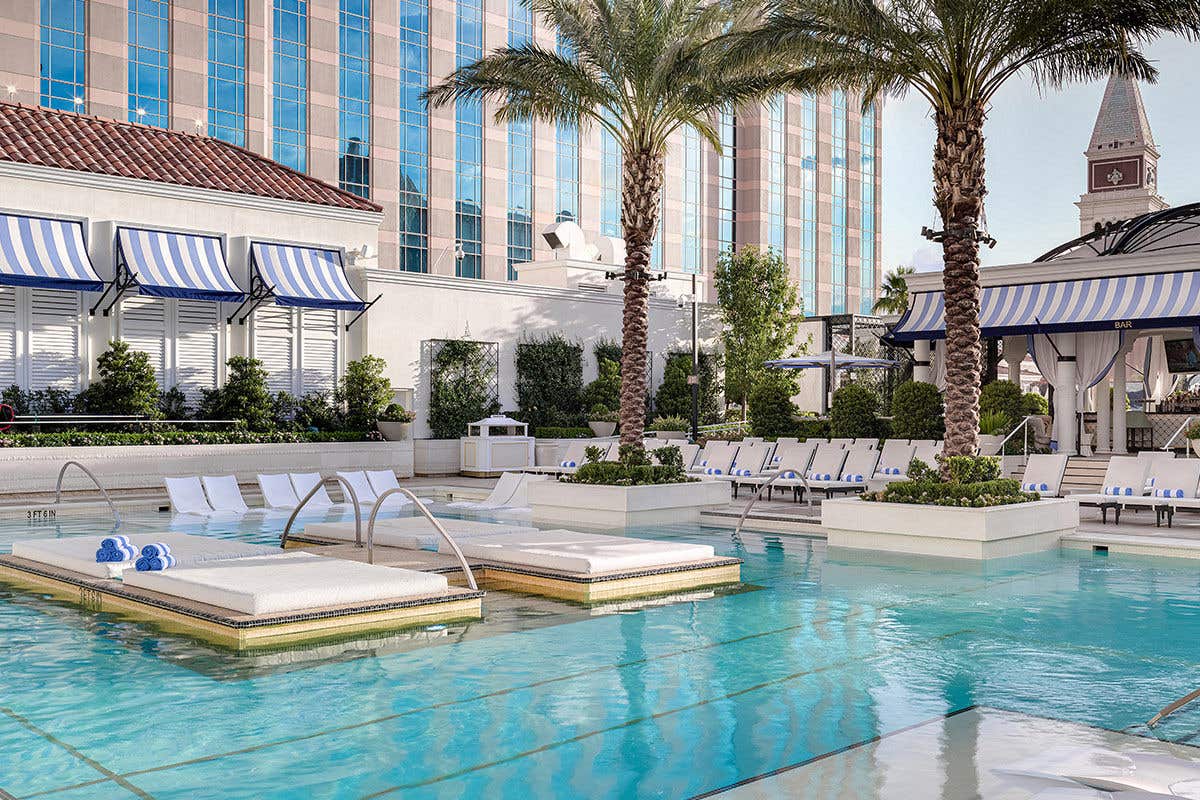 Island daybeds surrounded by pristine pool waters on The Venetian pool deck with palm trees, poolside lounge chairs, and the resort in the background.