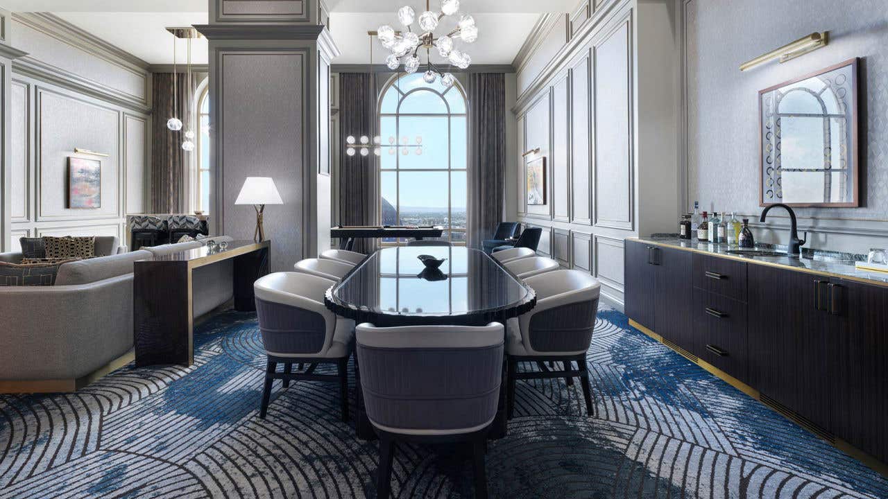 A hotel living room with a long oval dining table with eight chairs, a wet bar to it’s right, a couch to it’s left, a pool table behind it, and a tall arched window.