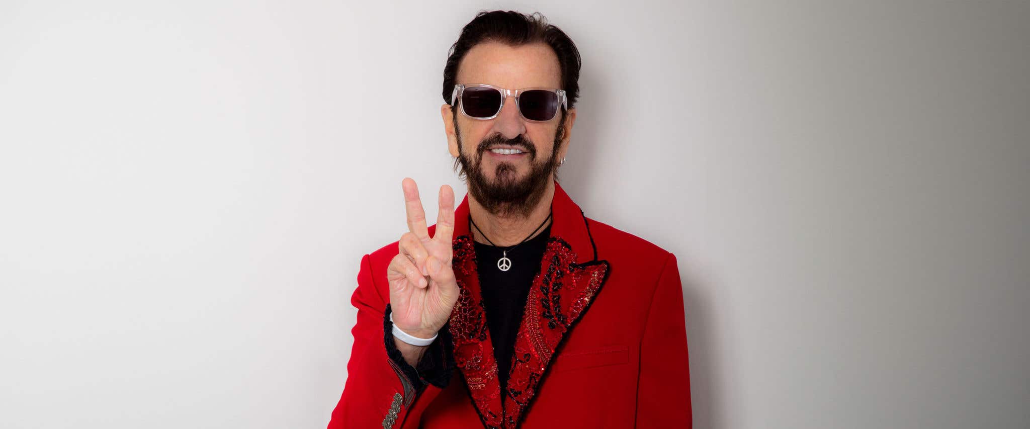 Man in a red jacket and sunglasses making a peace sign with his hand against a white background.