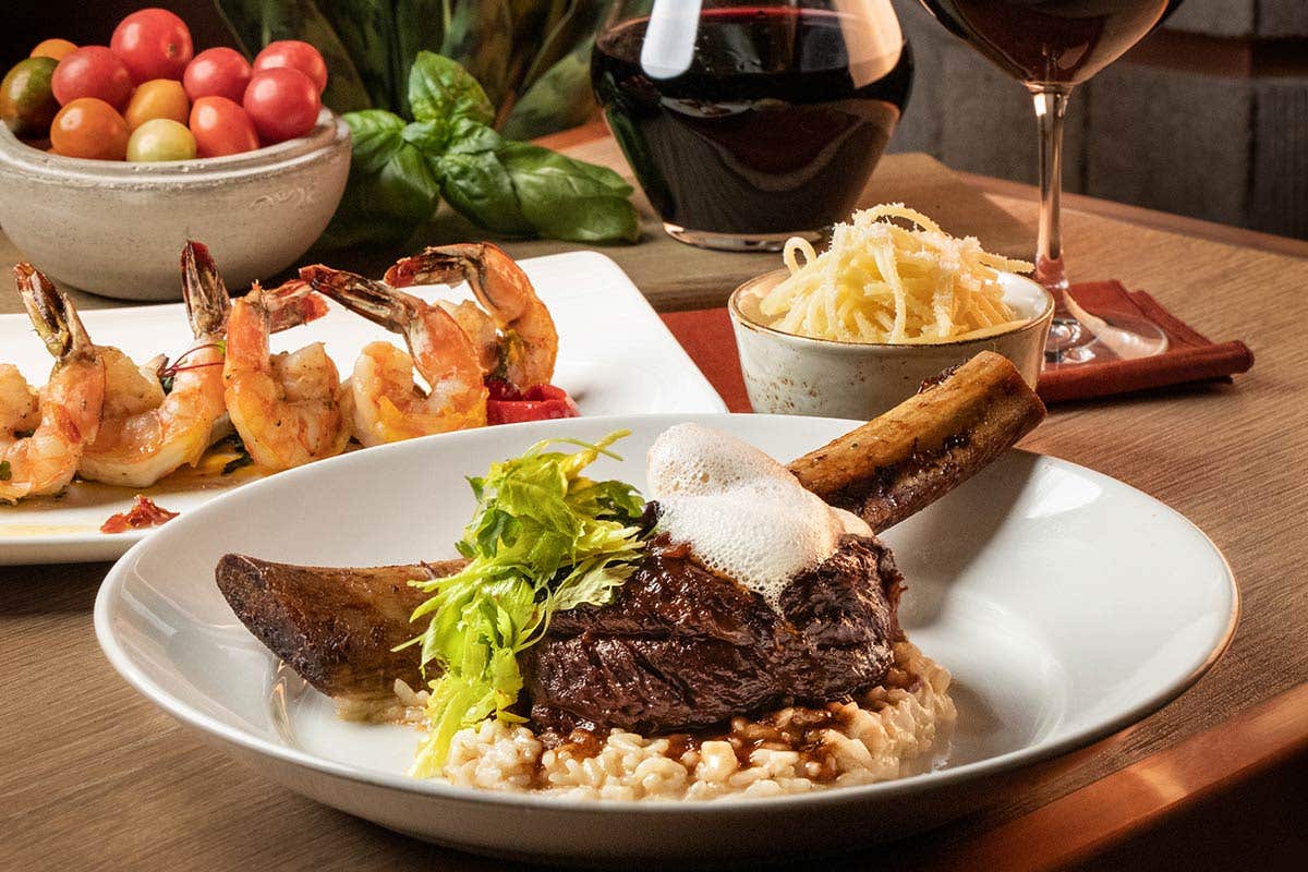 A plate featuring beef short rib on risotto, with a plate of shrimp, a bowl of spaghetti, tomatoes, basil, and wine in the background