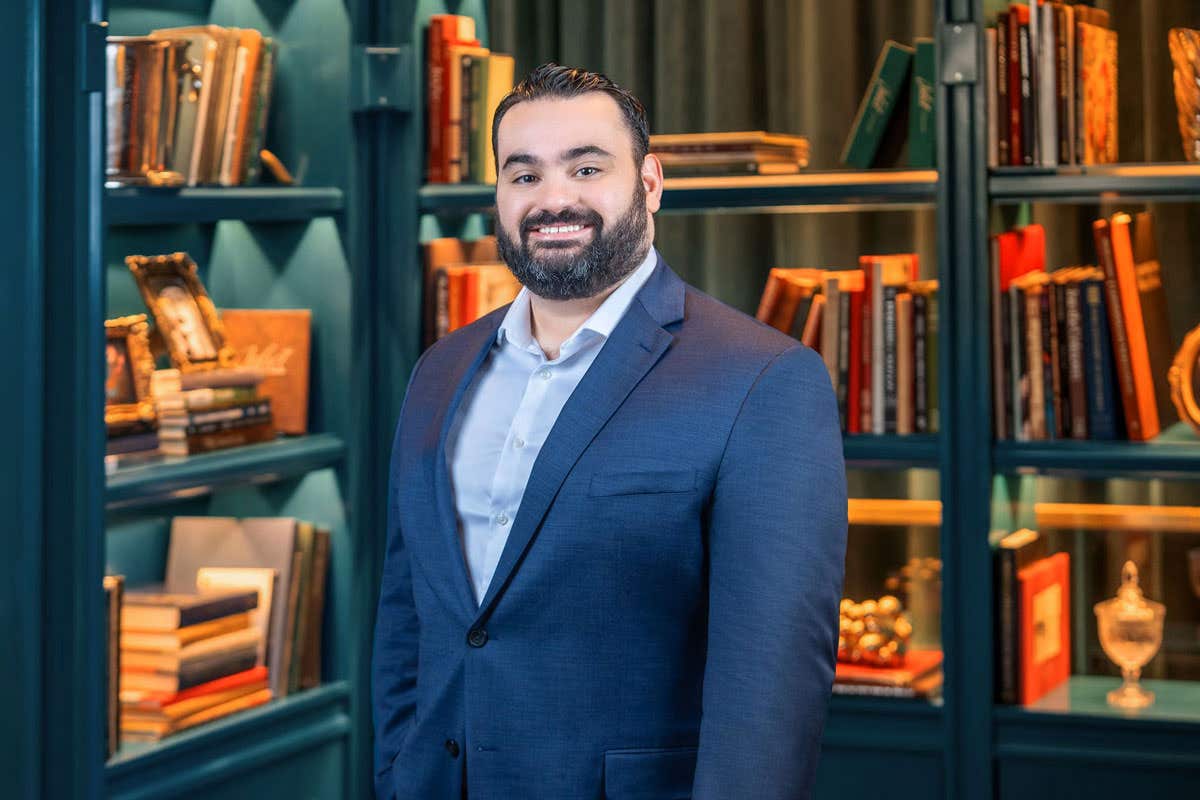 Man with short dark hair and a full beard, wearing a navy suit and light blue shirt, smiling in front of bookshelves in a warmly lit library setting.