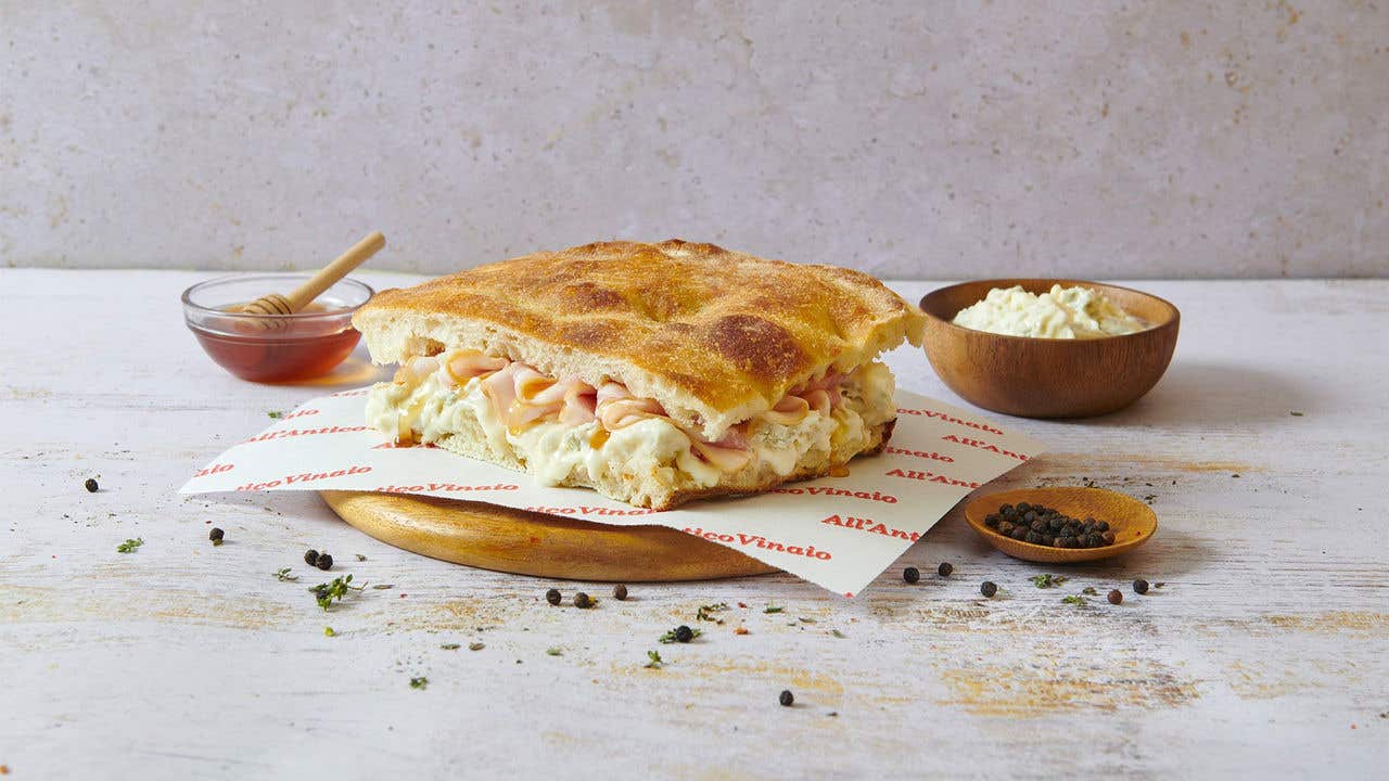 Panini sandwich with cheese and turkey on a round wood plate surrounded by a bowl of honey, parmesan cheese and peppercorn.
