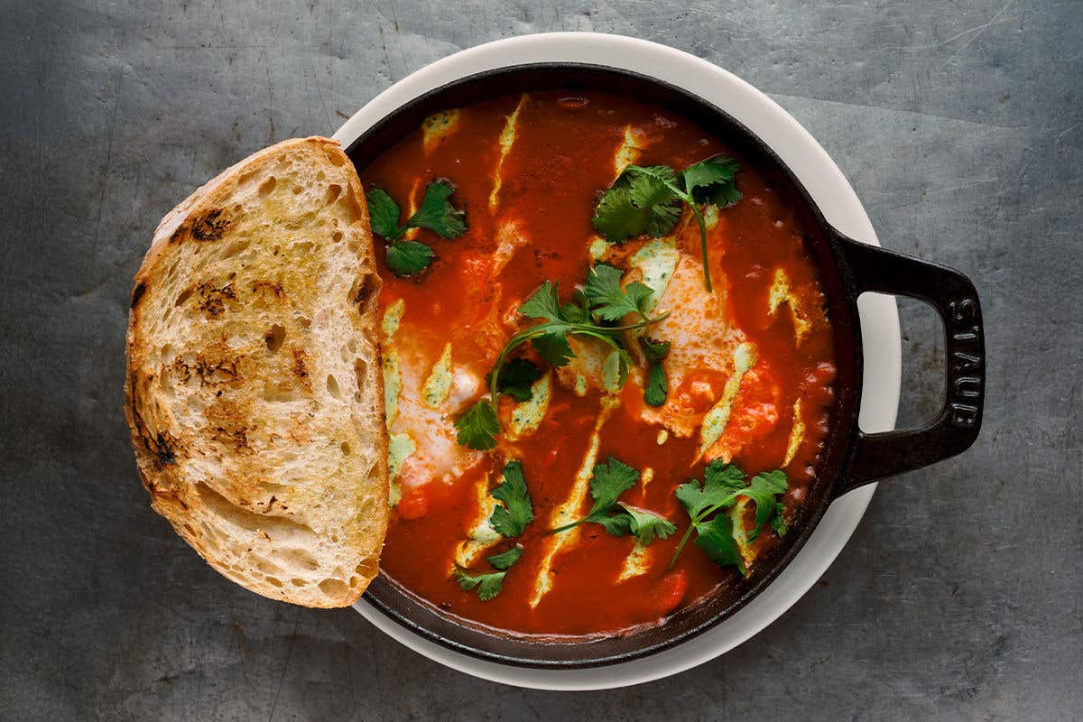 Baked eggs covered in tomato sauce in a cast iron bowl, with basil on top and a piece of toast on the side. 