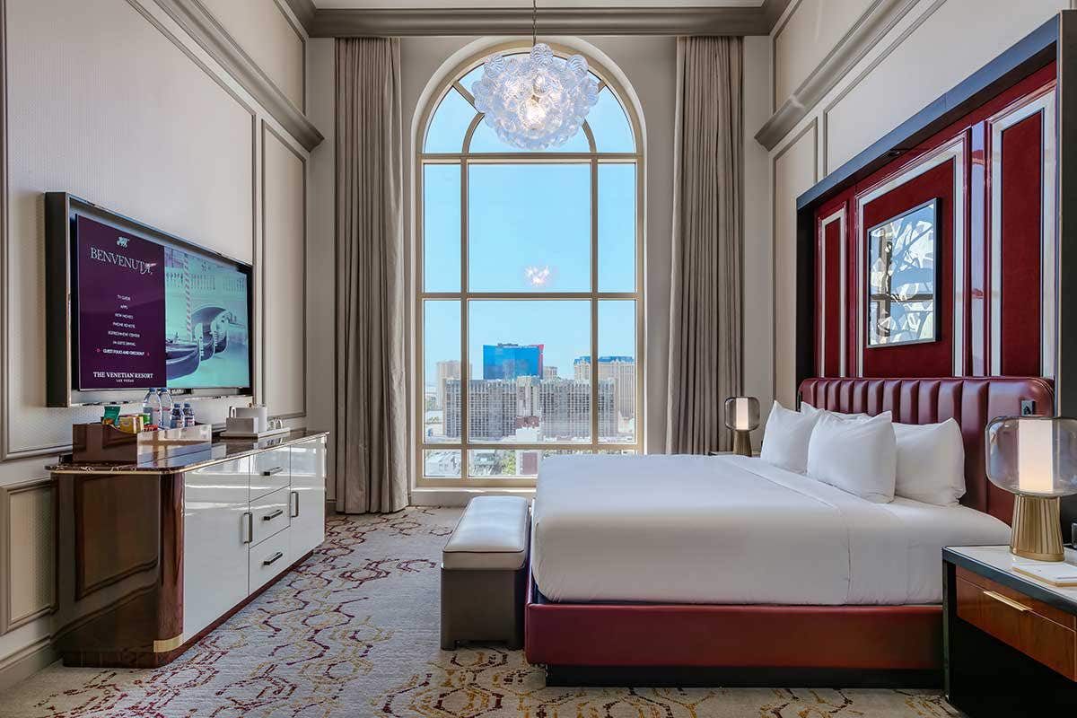 A hotel room with a king bed on the right, credenza and TV on the left, a chandelier above, and a large arched window straight ahead looking onto the Las Vegas Strip. 