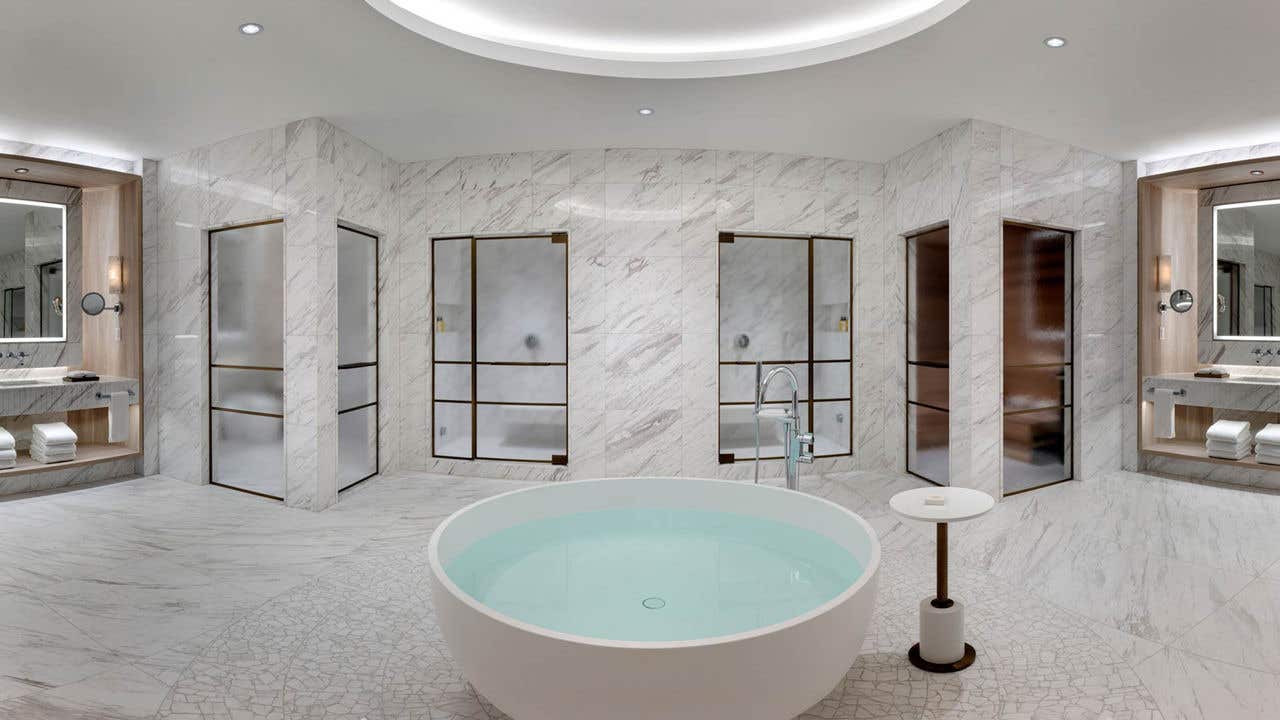 A large white marble bathroom with a round free-standing bathtub in the middle, two sink vanities on either side, and four frosted glass doors leading to a water closet, a shower, and a sauna.