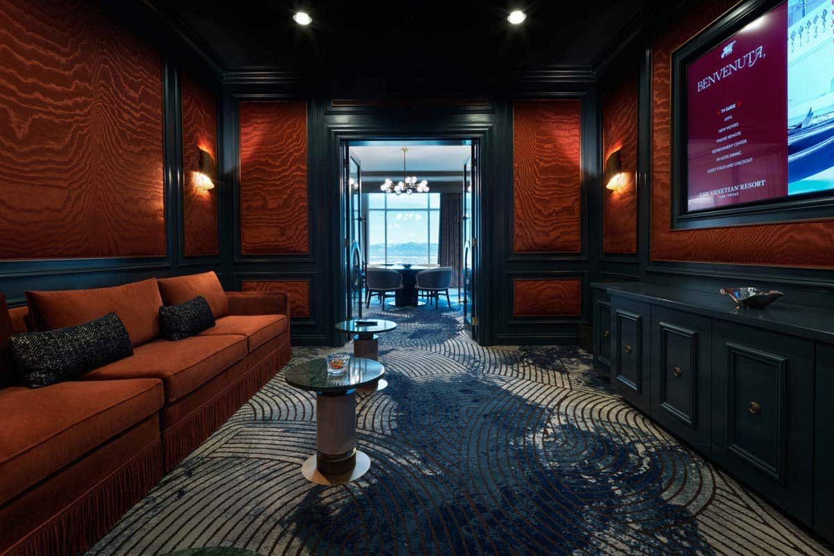 A hotel movie theater room with a long couch on the left, a large wall-mounted TV on the right, and a set of doors in the middle leading to a living room.