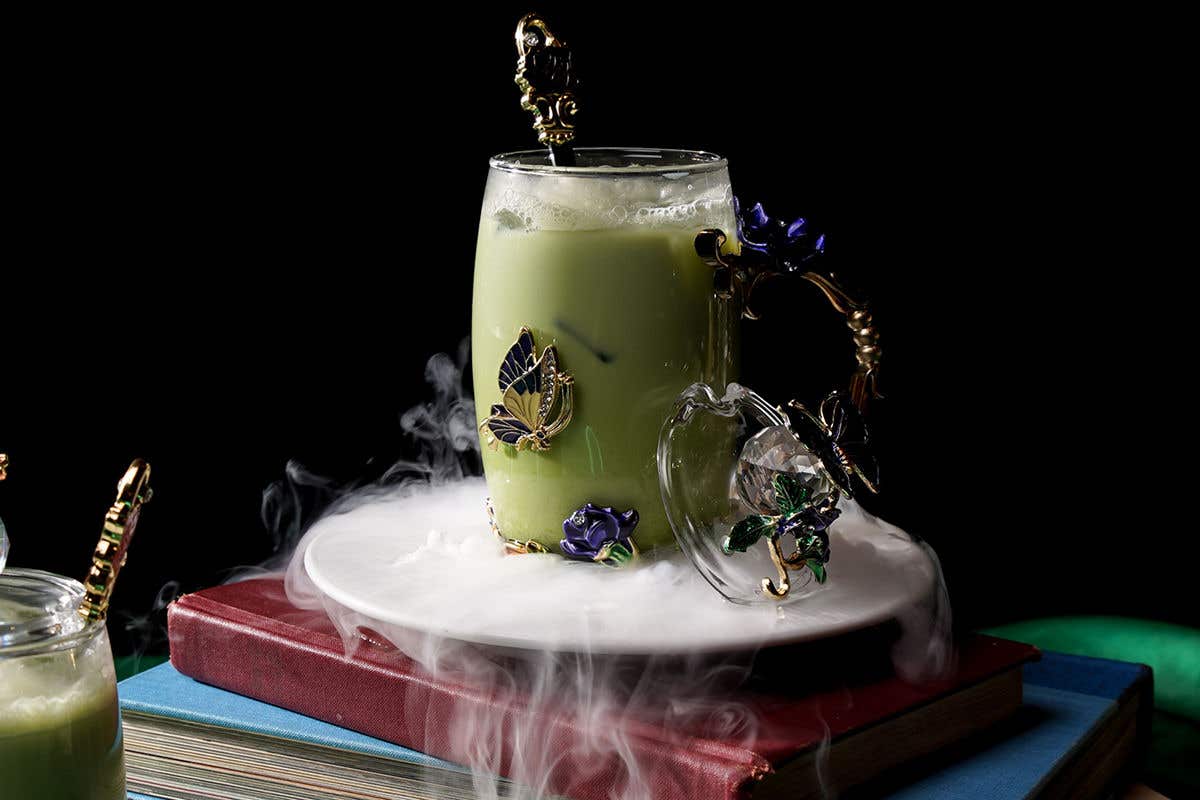 A green drink in an ornate glass cup, on top of a white plate with mist flowing off of it, placed on top of books, against a dark black background