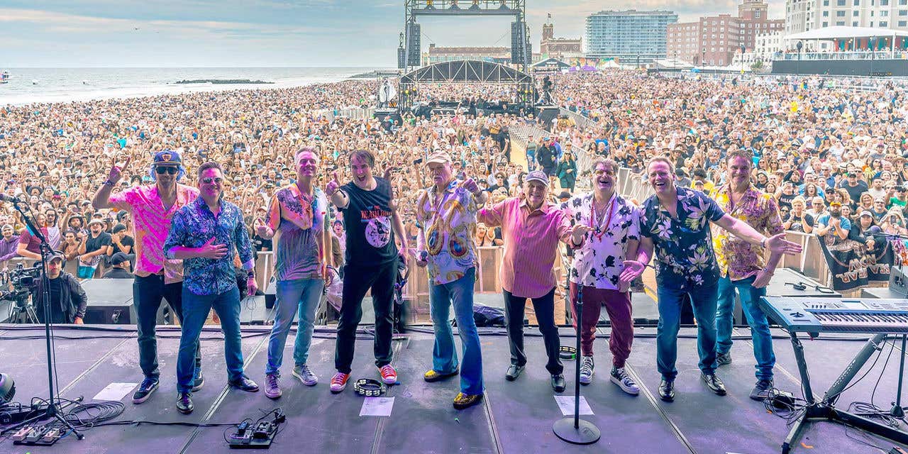 The Beach Boys, standing on stage facing the camera, with a large crowd of concert goers behind them.