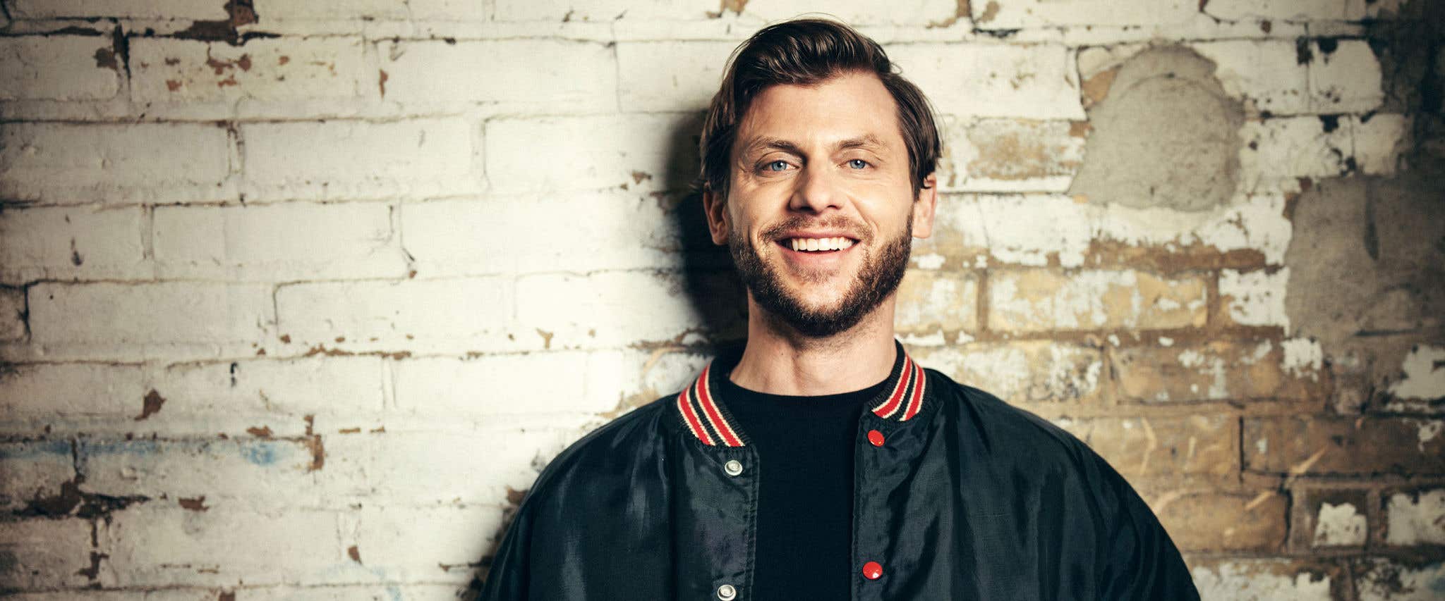 Charlie Berens in a black shirt and black jacket with red buttons smiling at the camera standing in front of a brick wall. 