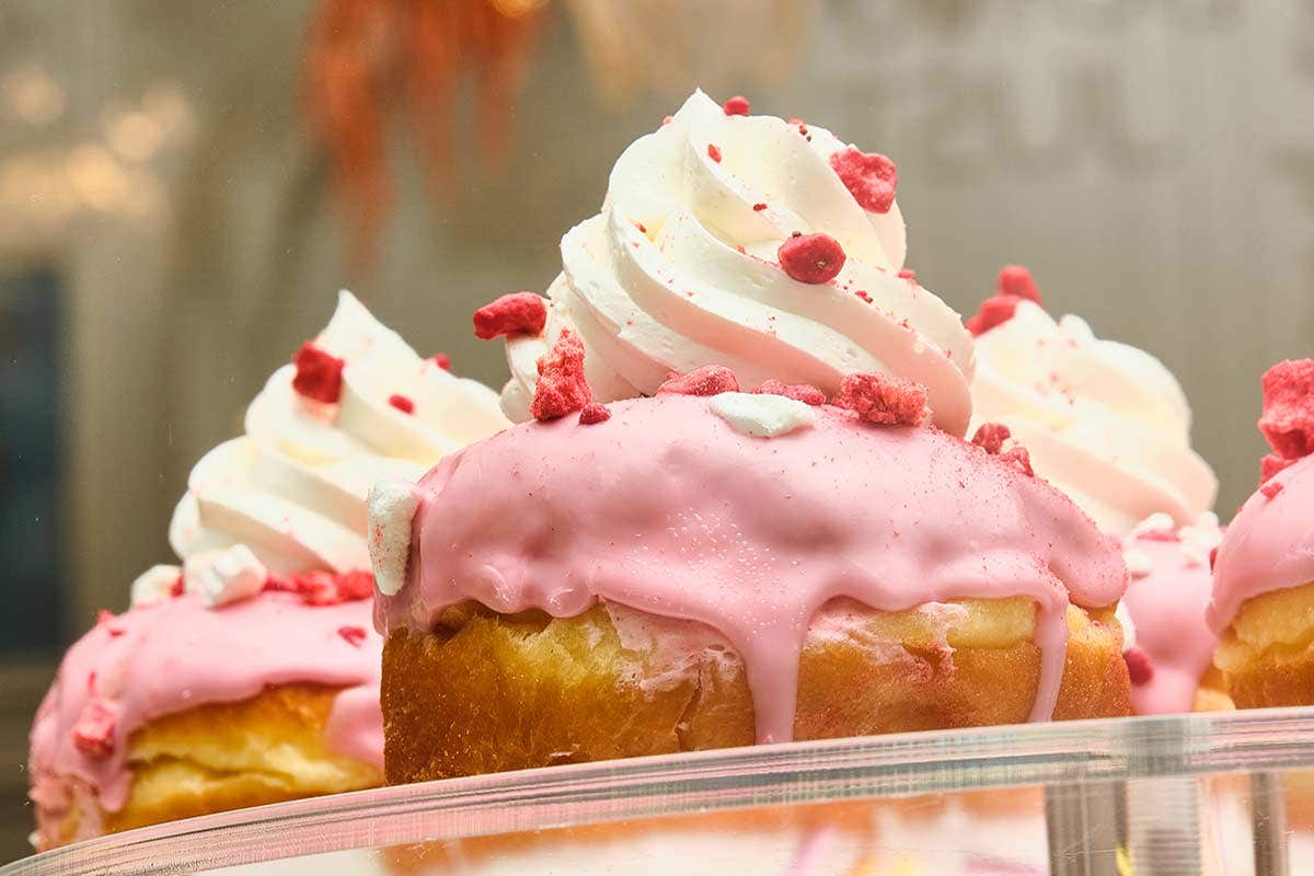 Close-up of pastries topped with pink icing, whipped cream, and red sprinkles displayed on a glass shelf.