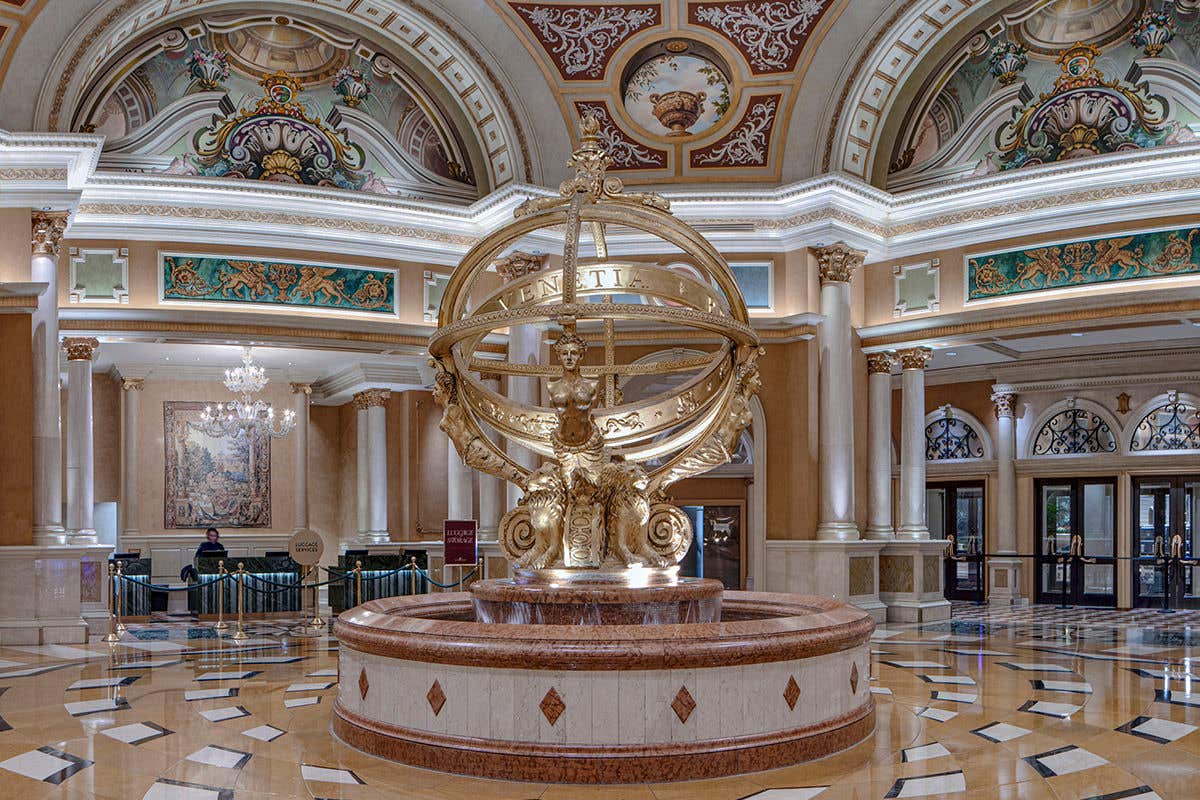 The Venetian lobby with a golden statue of lions and mermaids holding up interlocking golden rings that form a sphere. Grand columns and Renaissance style art adorns the walls, with the Luggage desk and entrance visible in the background. 