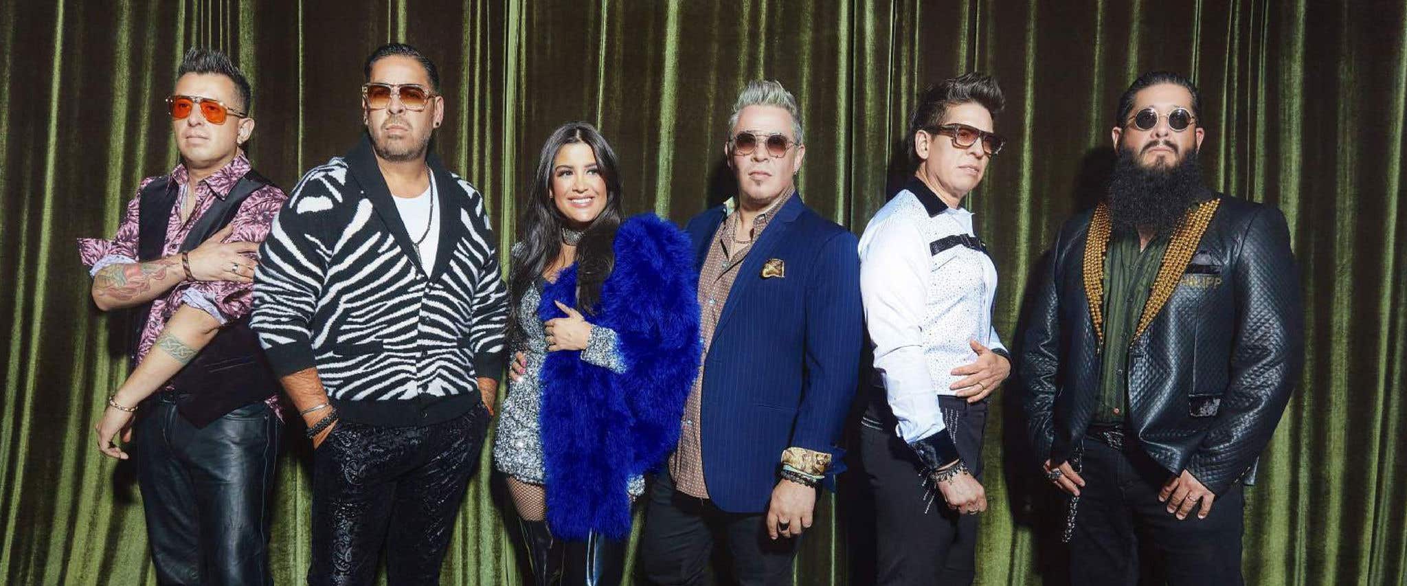 A group of six people standing in front of a green curtain, wearing stylish and colorful outfits.